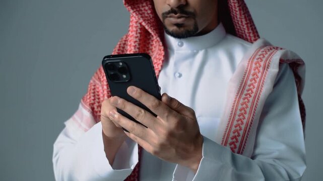 Close-up of a man in traditional Saudi attire using a smartphone against a gray background.