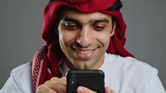 Smiling Arab man in traditional red headscarf looking at smartphone on gray background