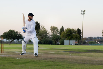 Obraz premium Male batsman in white kit standing at crease near stumps at dusk holding bat, copy space