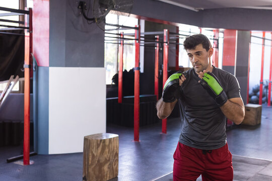 Man boxing in commercial gym wearing green-and-black gloves, gray shirt, red shorts, copy space