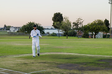 Obraz premium Male cricketer wearing white kit and pads standing on worn pitch at dusk holding wooden bat
