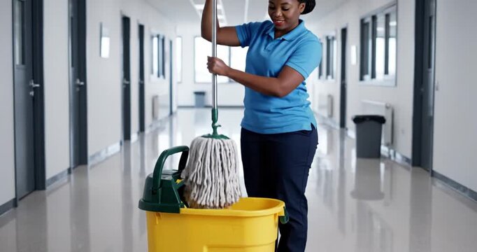 A smiling, professional facilities worker is diligently wringing out a wet string mop in a bright yellow bucket with a green wringer, situated in a clean, well-lit modern hallway. The reflective floor