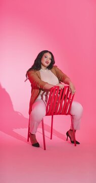 Vertical video: Mid-adult woman straddling red chair reacting to bright lights on pink backdrop
