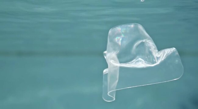 Translucent plastic debris, shimmering with light, gently floats in crystal clear blue underwater environment.