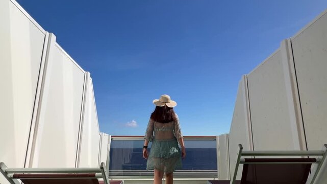 Latin american tourist female walking barefoot on a cruise ship deck, wearing a sunhat and sheer cover-up, enjoying a relaxing vacation and the ocean view under a clear blue sky.