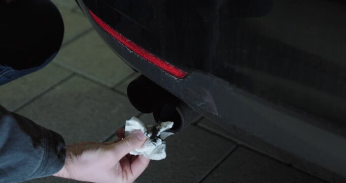 Close-up of a man wiping a car exhaust pipe with a tissue to check for soot or diesel particulate filter issues.