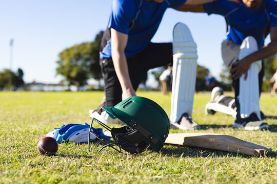 Green batting helmet is resting on grassy cricket pitch alongside gloves, ball, bat