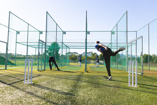 Bowler bowling in netted lane with metal poles while batsman preparing shot next to stumps on turf