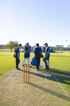 Cricket players standing on pitch around stumps in blue and black jerseys under afternoon sun