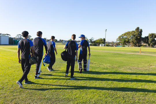 Cricket players walking across suburban grassy pitch, carrying bats and pads