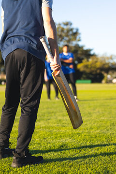 Batsman standing on lush cricket field with wooden bat facing distant fielders under blue sky