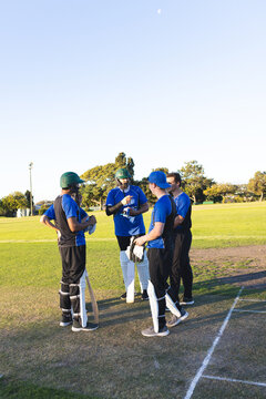 Five cricket players standing on sunlit grass pitch next to boundary line, holding bats and gloves