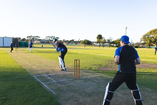 Cricket match unfolding at suburban ground, stumps at crease and ball soaring toward fence