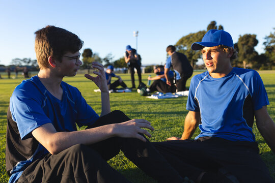 Teenage athletes are sitting on grass in blue jerseys and are talking under late afternoon sun