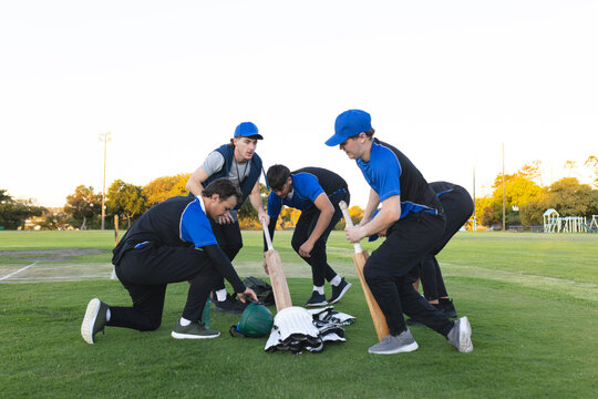 Five cricket players gathering at black kit bag, picking bats, helmets and pads on ground at dusk