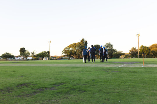 Group of male cricket players walking across grassy field next to stumps carrying bat
