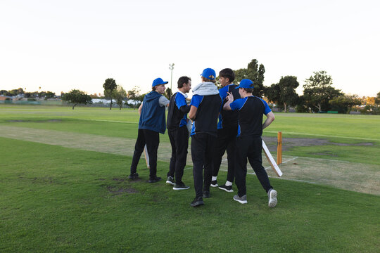 Six cricket players are celebrating in huddle next to wooden stumps on grass pitch under clear sky