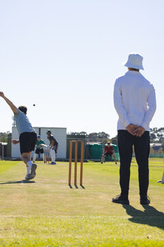 Bowler delivering ball on grass pitch, with stumps, batsman, keeper, fielders under clear sky