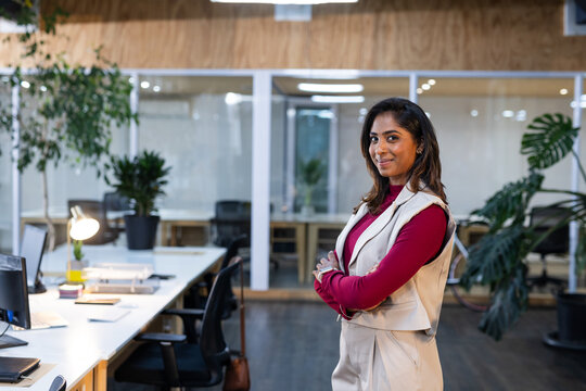 Indian woman in business casual crossing arms in coworking office by desk with handbag, copy space
