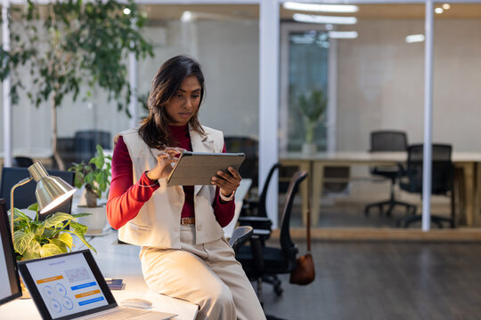 Indian businesswoman sitting on desk interacting with tablet at office with lamp and laptop charts