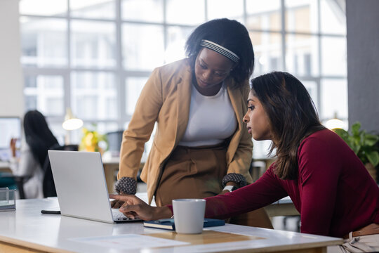 Diverse female coworkers collaborating at office desk using laptop, coffee mug and notebook