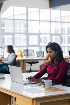 Diverse female coworkers in business attire typing on laptops at open-plan office desks, copy space
