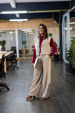 Asian Indian woman holding laptop while standing in open plan office with ergonomic chairs, plants