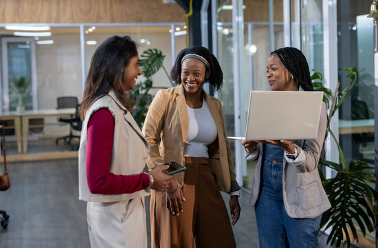 Diverse female coworkers standing in glass-walled office corridor holding laptop and smartphone