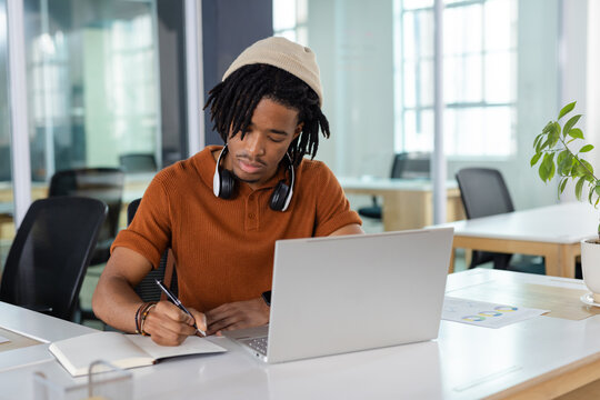African American man wearing headphones sitting at office desk writing in notebook with laptop