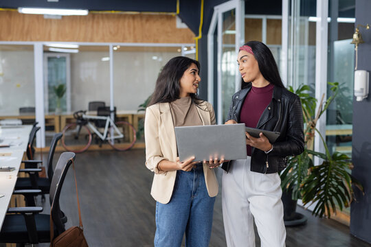 Diverse female coworkers wearing blazers collaborating on laptop and tablet by bicycle at office