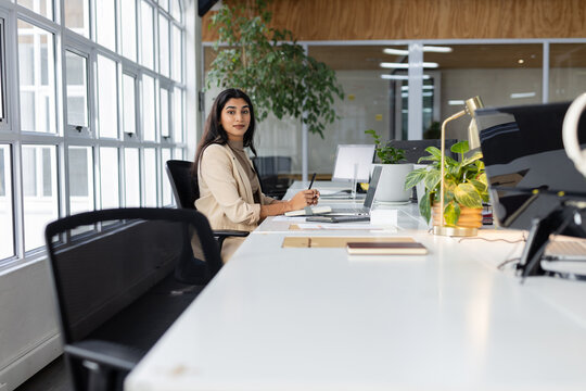 Indian woman working on laptop and monitor at office workstation with notebook lamp and plants