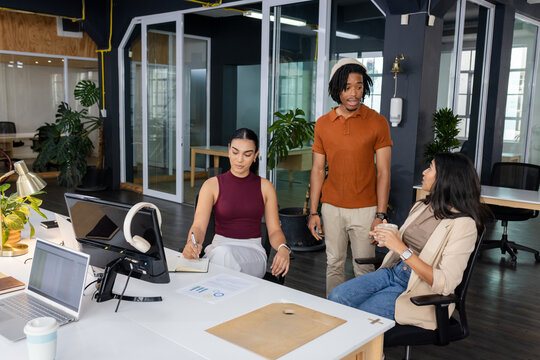 Diverse coworkers reviewing printed charts at open-plan desk while using laptop and sipping coffee