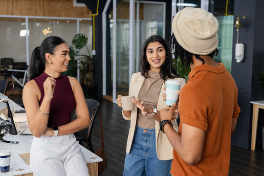 Diverse coworkers chatting while holding coffee mugs near workstation in modern open-plan office