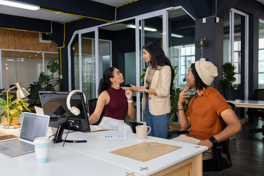 Diverse coworkers in office attire discussing project around laptops with coffee mugs at white desk