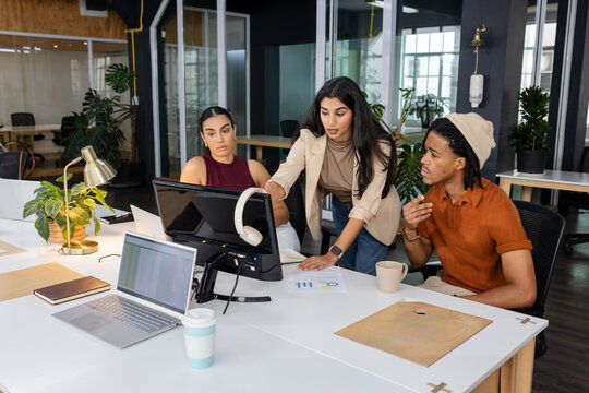Diverse coworkers analyzing data on desktop monitor with printed charts at coworking office