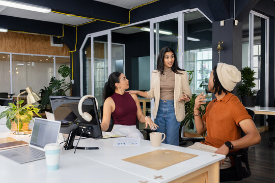Diverse coworkers gathering around desk in open office discussing project with laptops and charts