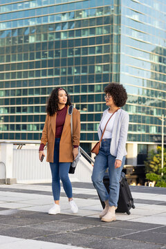Diverse female friends walking on rooftop path wearing blazers pulling suitcase, with smartphone
