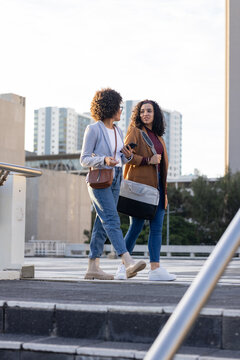 African american female friends walking on parking deck by handrail holding smartphone, bags