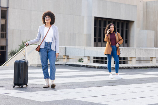 Female friends standing on terrace pulling suitcase, carrying bags, talking on smartphone