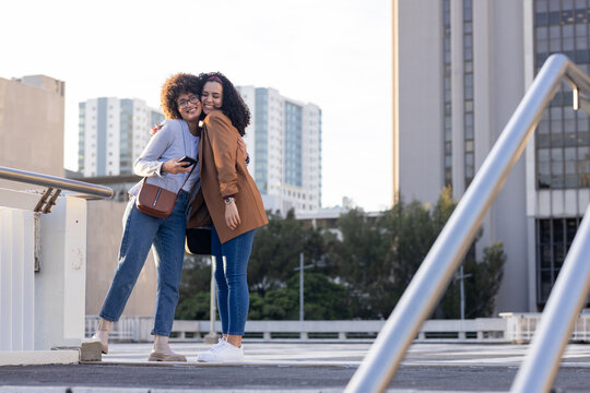 Female friends hugging, leaning against metal railing on rooftop deck holding smartphone