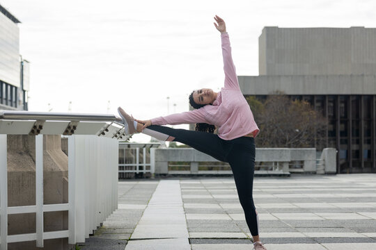 Woman performing high side stretch using metal railing on urban terrace in pink hoodie