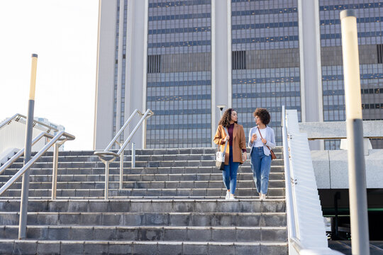 Diverse female friends walking down stairs at office plaza holding coffee cups, bags, copy space