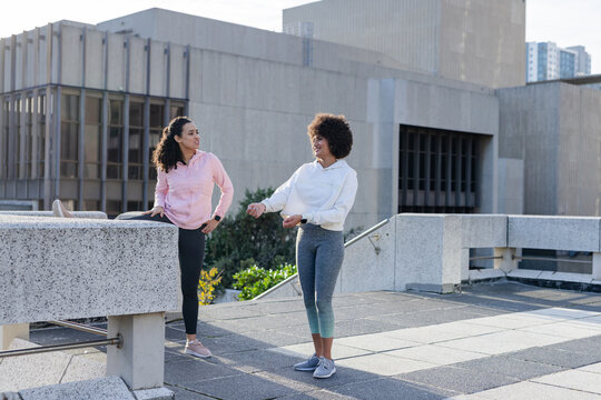 Diverse female friends stretching hamstrings on concrete bench on urban terrace in hoodies