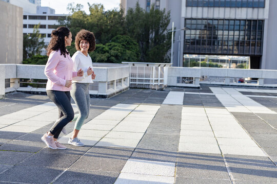Two joggers running across tiled rooftop terrace overlooking trees, office building facade