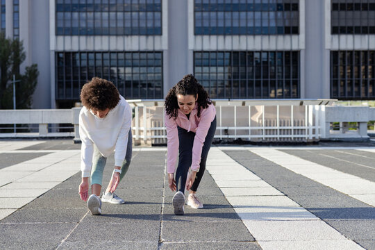 Diverse female friends stretching hamstrings on rooftop by railing in sportswear in trackers