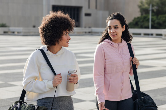 Diverse female friends strolling in hoodies across striped plaza with water bottle, duffel bags