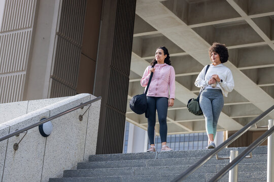 Diverse female friends in sportswear pausing on concrete stairs, gym bags, copy space
