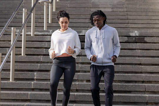 Diverse couple descending plaza stairs wearing hoodies gripping handrail, checking smartwatch
