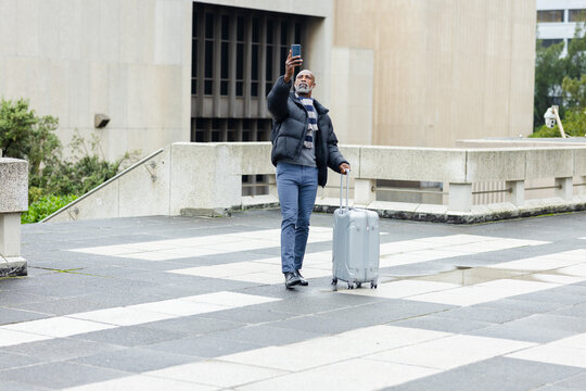 African american man standing in city square holding smartphone aloft and pulling rolling suitcase