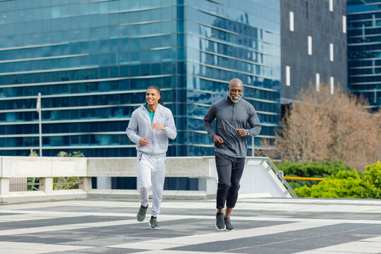 Diverse male friends running with braids and watch jogging on urban plaza with concrete railing